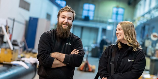 Två svartklädda, glada personer på en industriarbetsplats.