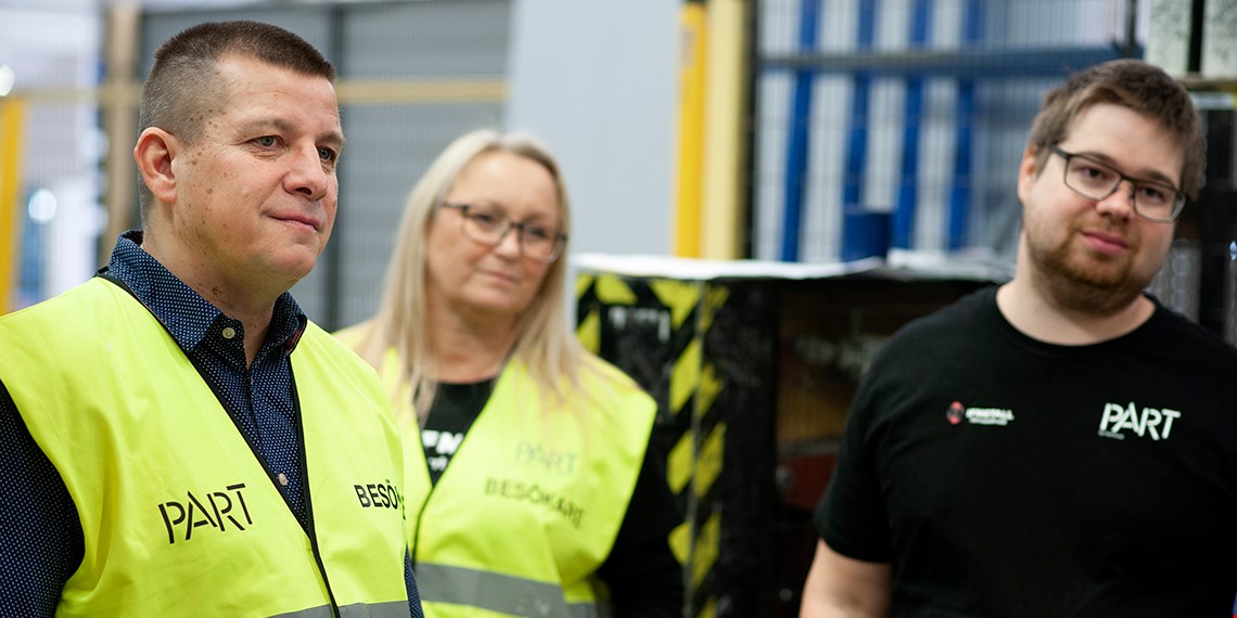 Arbetsplatsbesök på industri. Två besökare i gula västar och en person i svart t-shirt.