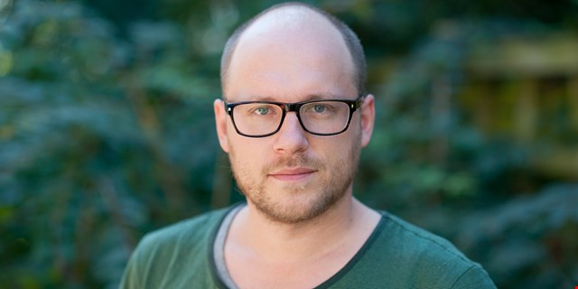 Person med glasögon och grön tröja framför suddig, grön bakgrund.