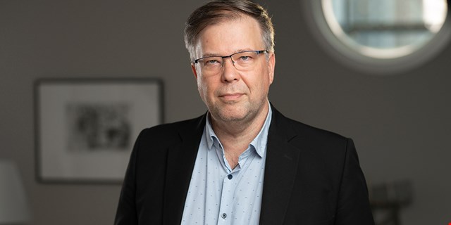 Porträtt av person i kavaj och blå skjorta mot mörk bakgrund