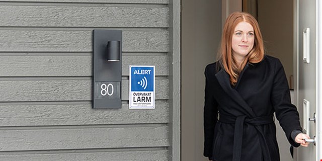 En kvinna öppnar dörren till sitt hus. På husväggen finns en skylt där det står Alert Alarm.